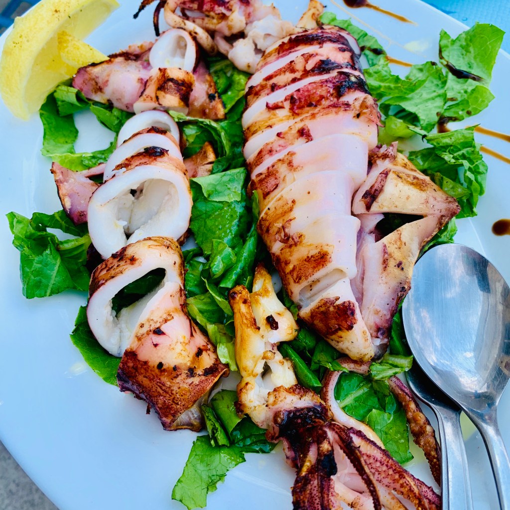 A plate of grilled squid on a bed of rocket with a lemon wedge, on a taverna table.