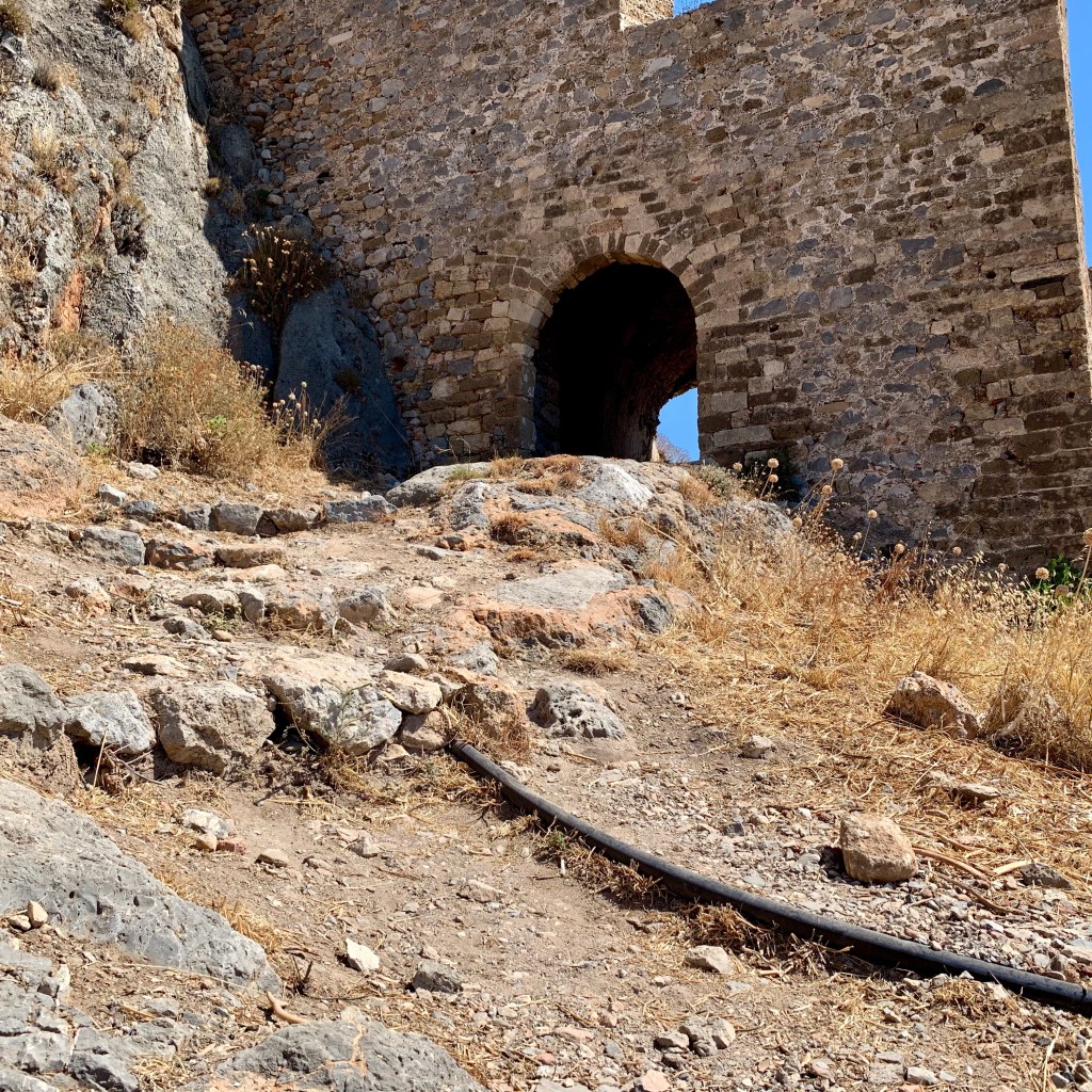 A stone archway in the fortress wall above Monemvasia, reached by a rough path climbing the hillside.