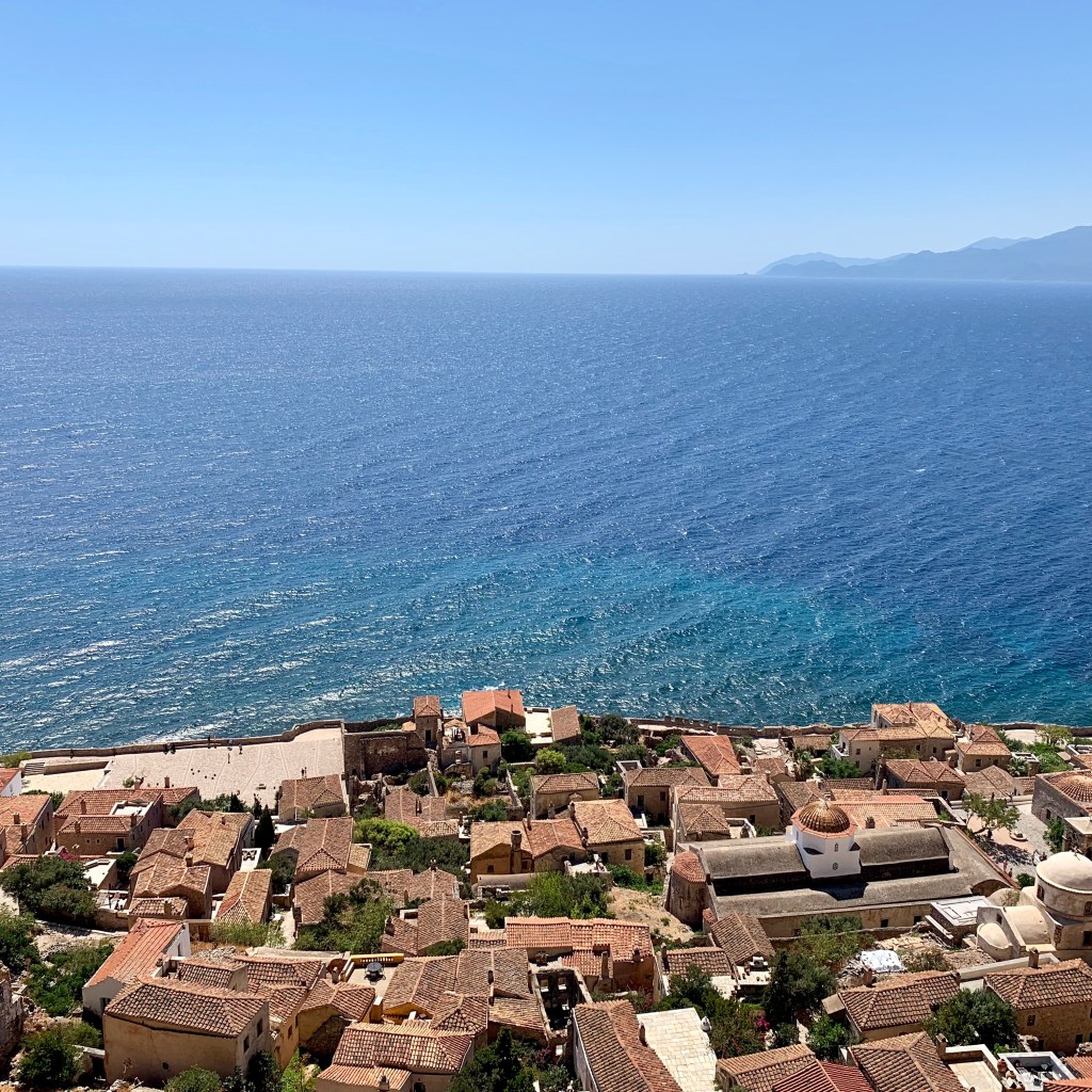 Looking down on the tiled roofs and domes of the lower town of Monemvasia, with open sea and a distant headland beyond.