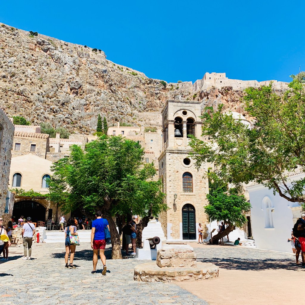 The main square of Monemvasia: a stone church and bell tower, plane trees, visitors on the flagstones, and the cliff face and upper fortress rising behind.