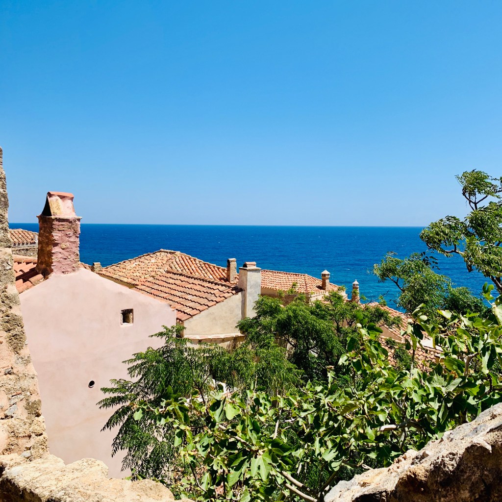 A view out to the sea over red-tiled roofs and a stone chimney, framed by a rough wall and green foliage.