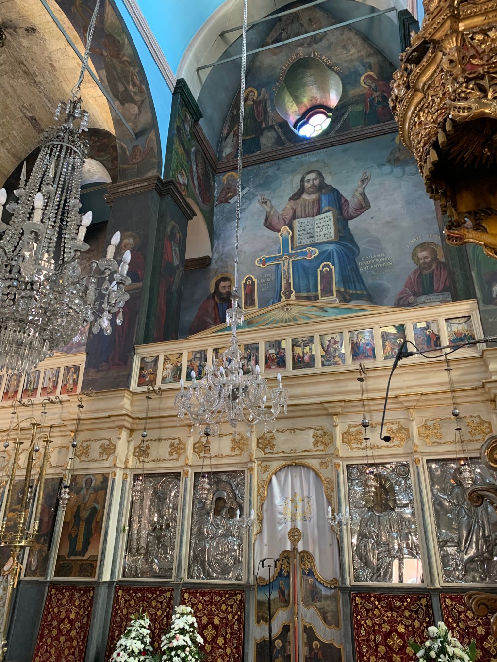 The interior of an Orthodox church in Chania: a tiered iconostasis of silver-clad icons below a painted Christ Pantocrator, with two large crystal chandeliers hanging in the nave and white flowers at the front.