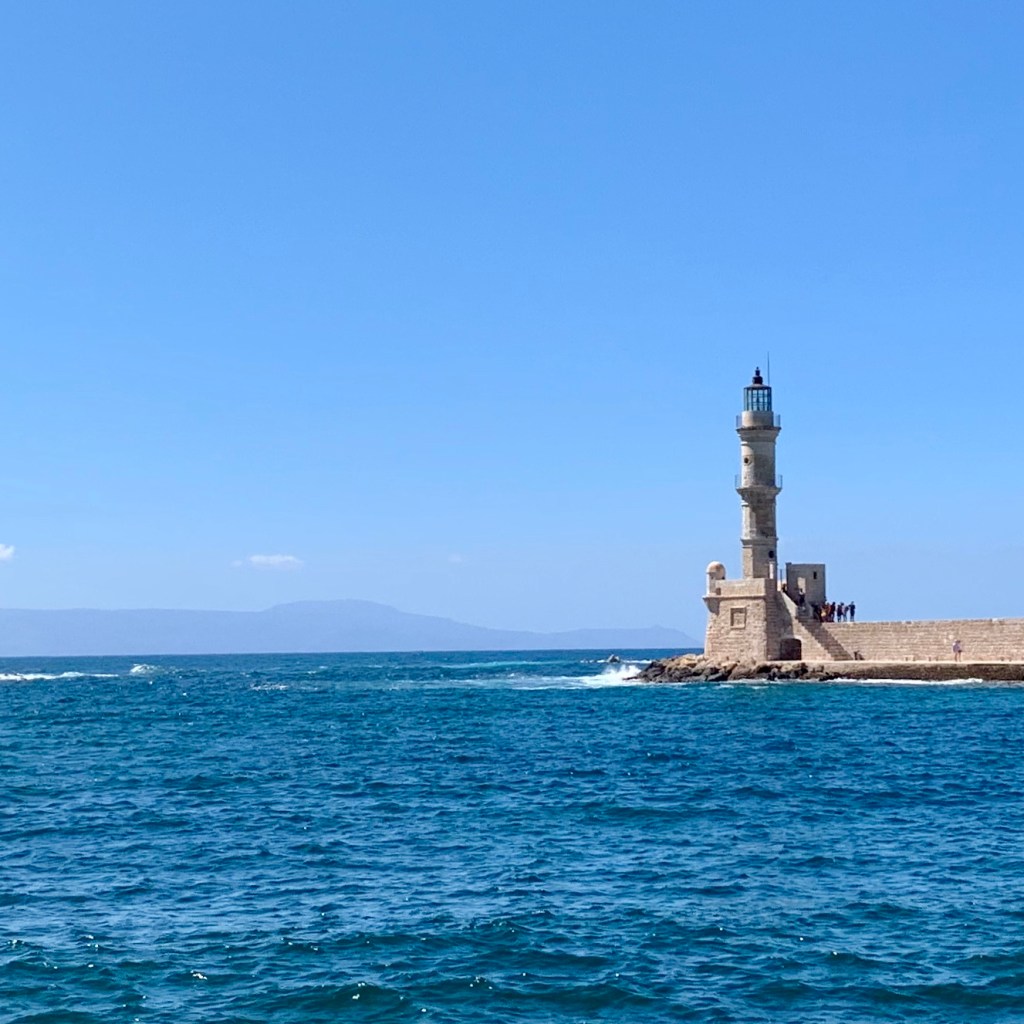 The Venetian harbour at Chania seen across flat blue water, with the stone mole running out to the domed Egyptian lighthouse and ochre waterfront buildings on the left.