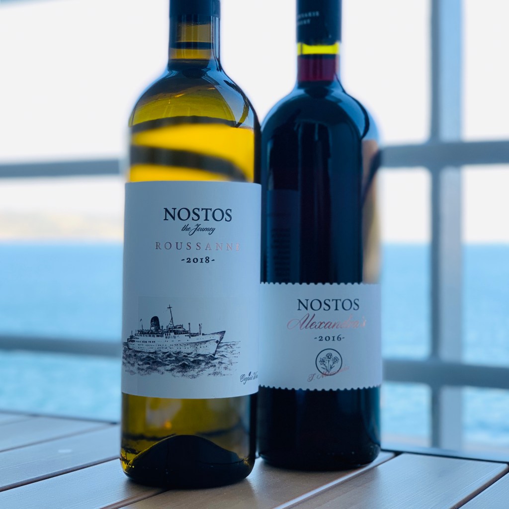 Two bottles of Nostos wine from the Manousakis winery standing on a teak table at the ship’s rail — a 2018 Roussanne in green glass and a 2016 Alexander’s red — with the Cretan coast behind them.