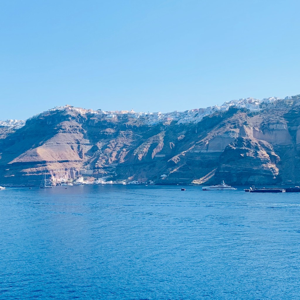 The caldera cliffs of Santorini seen from the water, the white buildings of Fira strung along the rim high above, ships lying at anchor below.