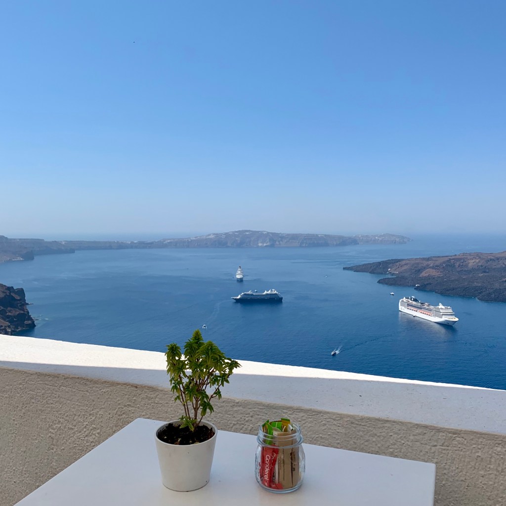 A whitewashed terrace with a small potted plant on a table, looking out across the caldera to two cruise ships at anchor and the volcanic islet beyond.