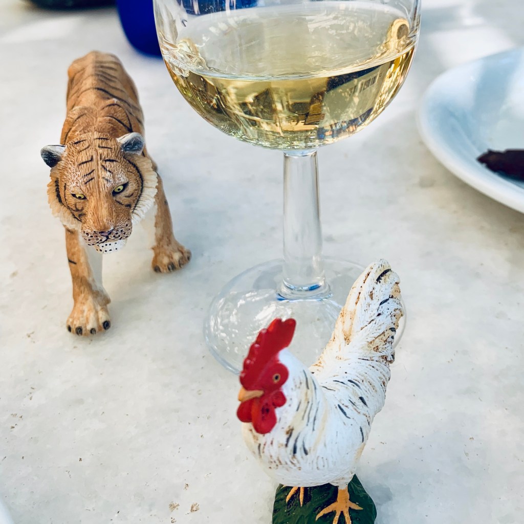 The tiger and rooster figurines standing on a restaurant table beside a glass of white wine and a plate of food.