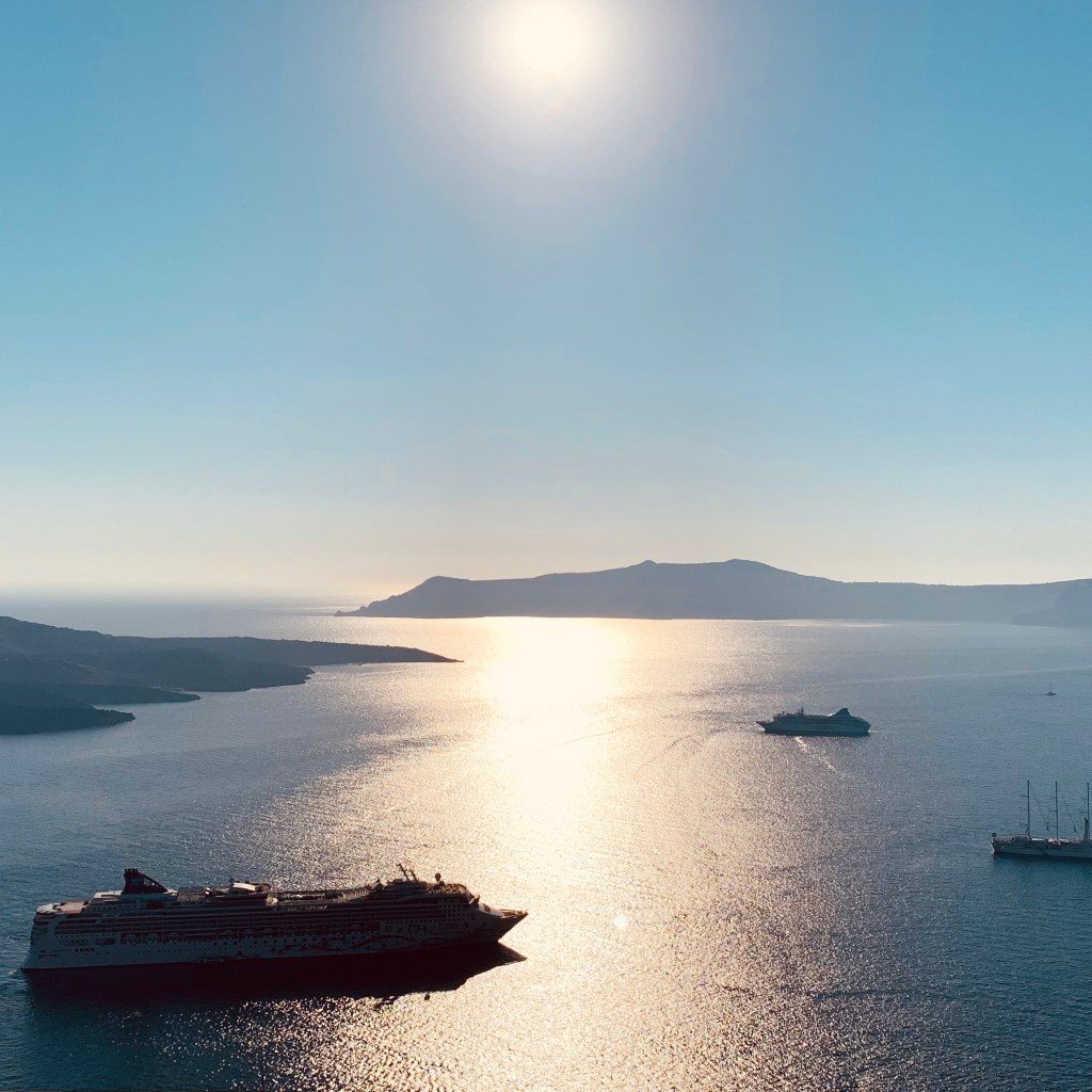 Late afternoon sun over the caldera, ships silhouetted on the bright water far below the terrace rail.