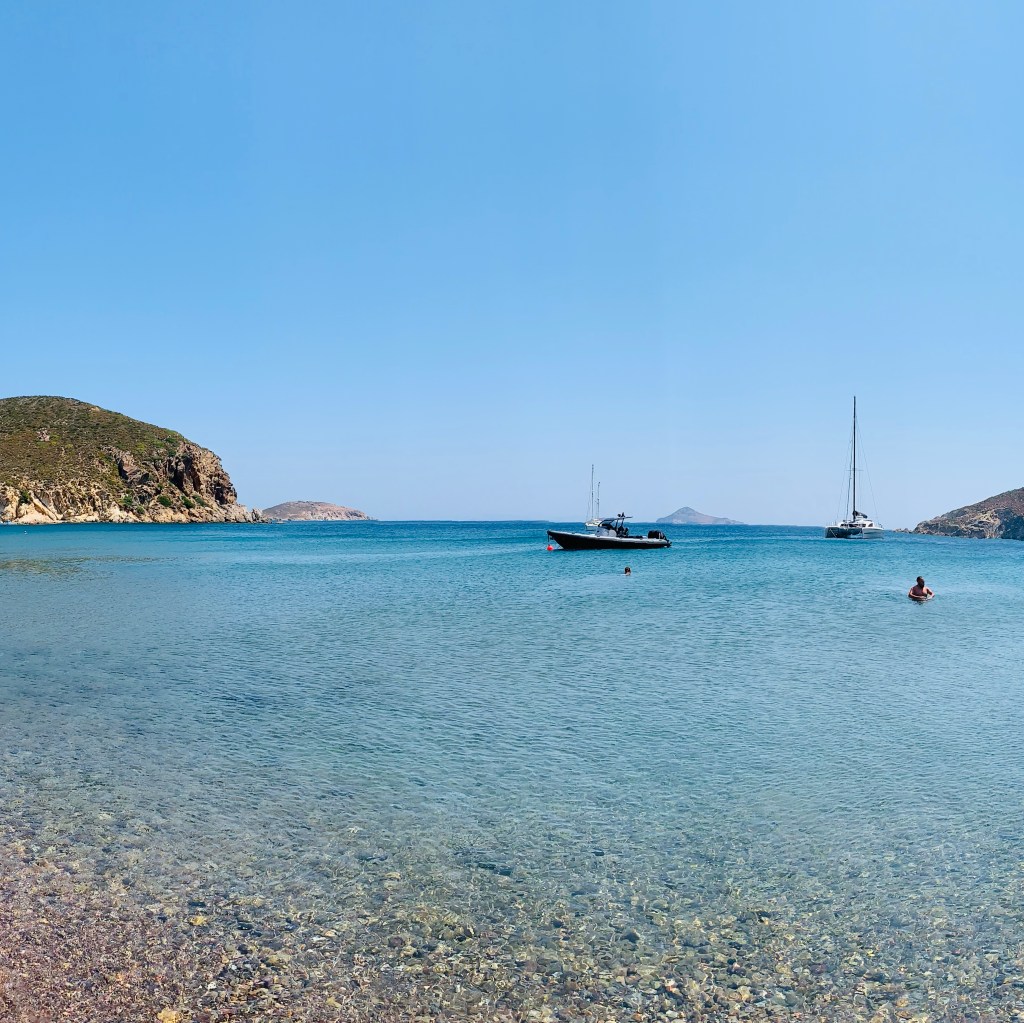 A shallow bay of very clear water with a pebbled shore, small boats and two yachts at anchor, and low headlands on either side.