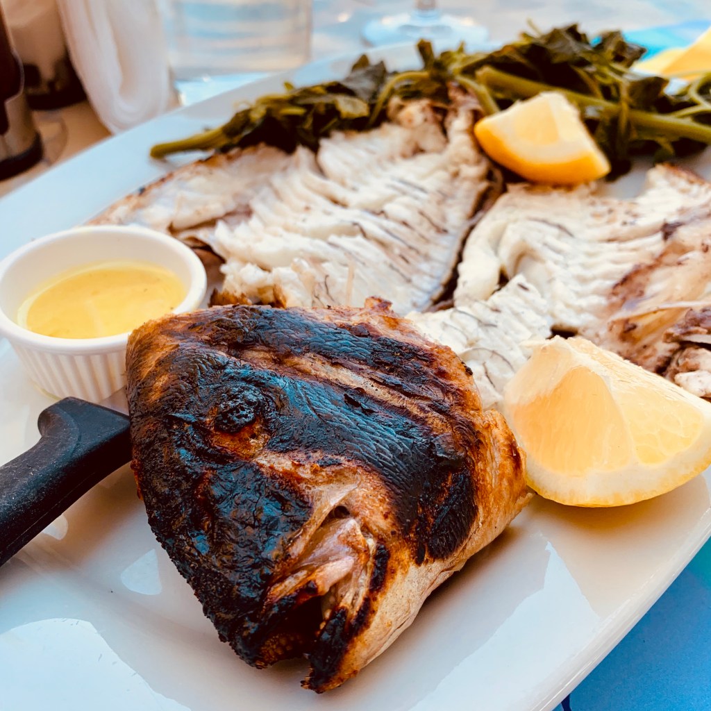 A whole grilled fish on a white plate with lemon wedges, a bowl of olive oil and a pile of wild greens.
