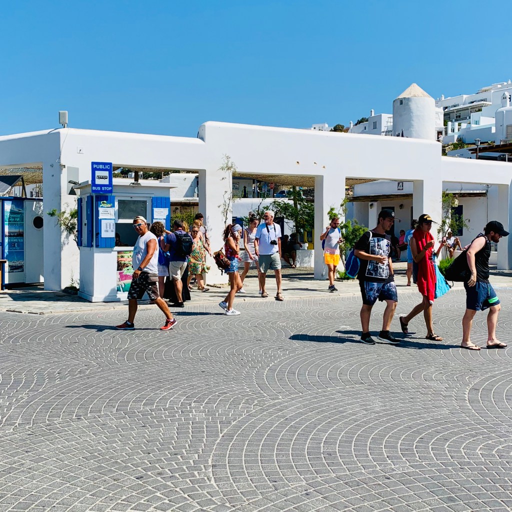 The white terminal building at the Mykonos tender pier, passengers crossing the patterned grey paving in front of it.
