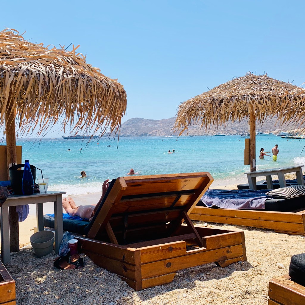 A beach of wooden loungers under thatched parasols, turquoise water and swimmers beyond.