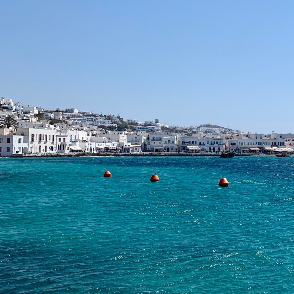 Mykonos town seen low across the water, a long line of white buildings against a bare hillside.