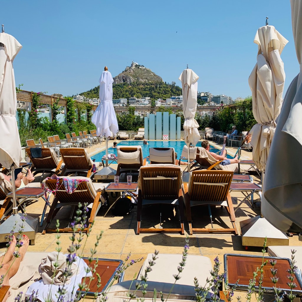 The hotel rooftop pool with teak loungers and furled white umbrellas, the cone of Lycabettus hill rising over the city beyond.