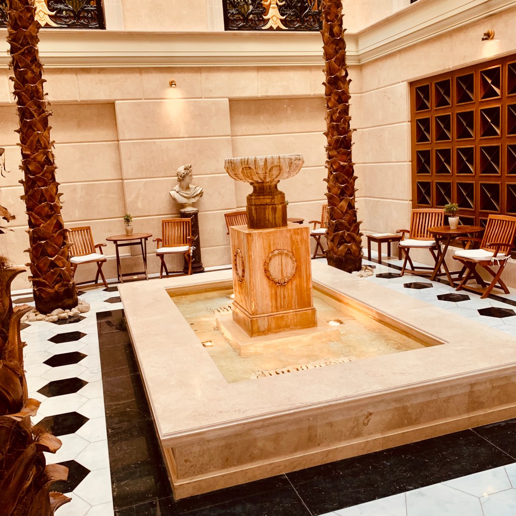 An interior courtyard with a carved marble fountain, tall palms in planters and a black and white tiled floor.