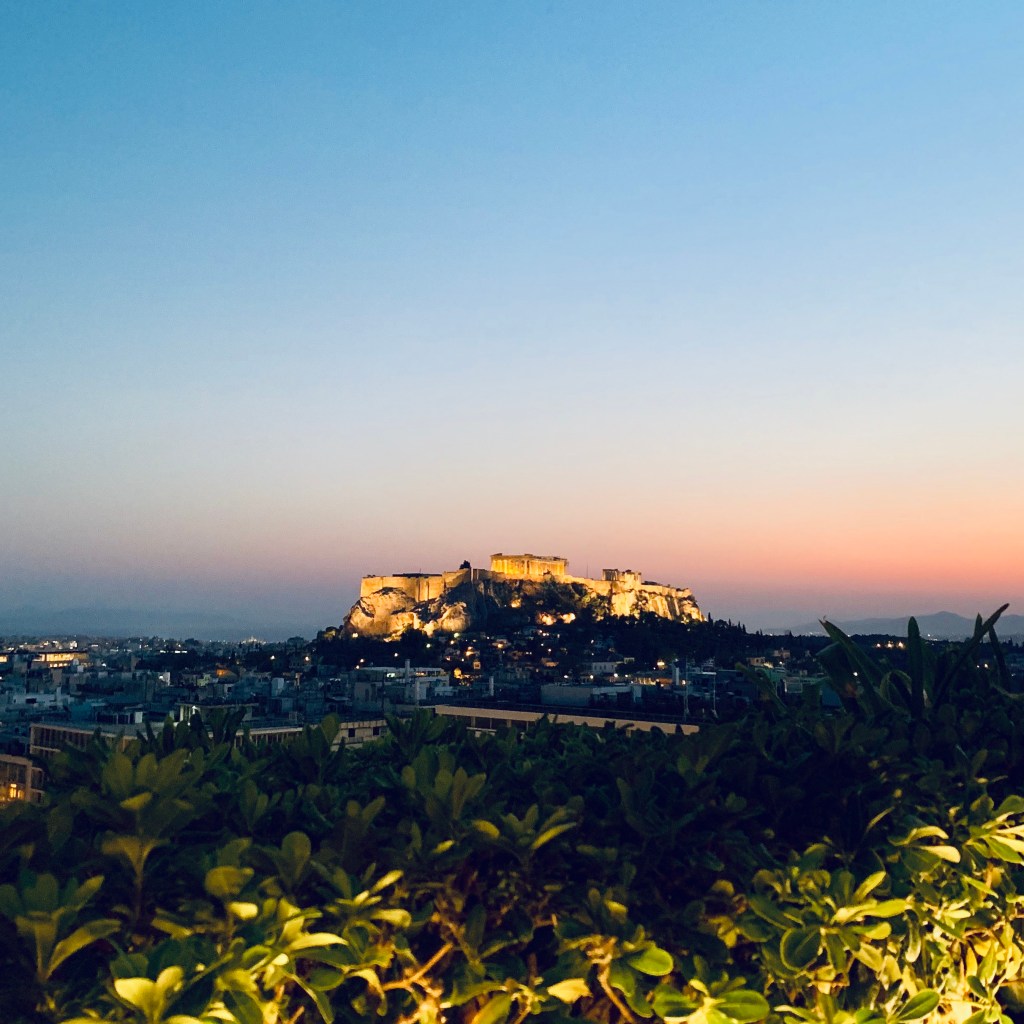 The Acropolis lit against a fading orange sunset, seen over the rooftops of Athens from the hotel terrace.