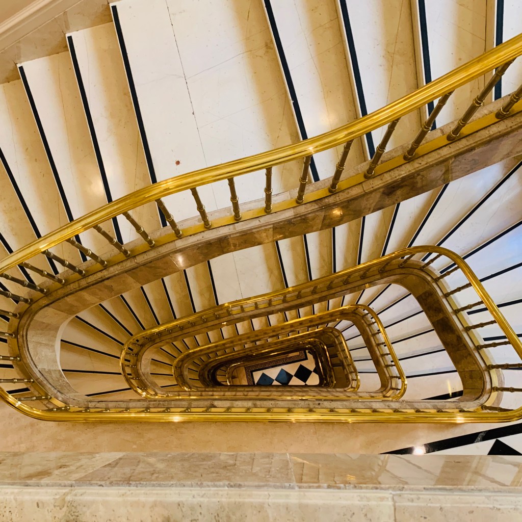 A stairwell seen from above, the marble steps and polished brass banister spiralling down through several floors.