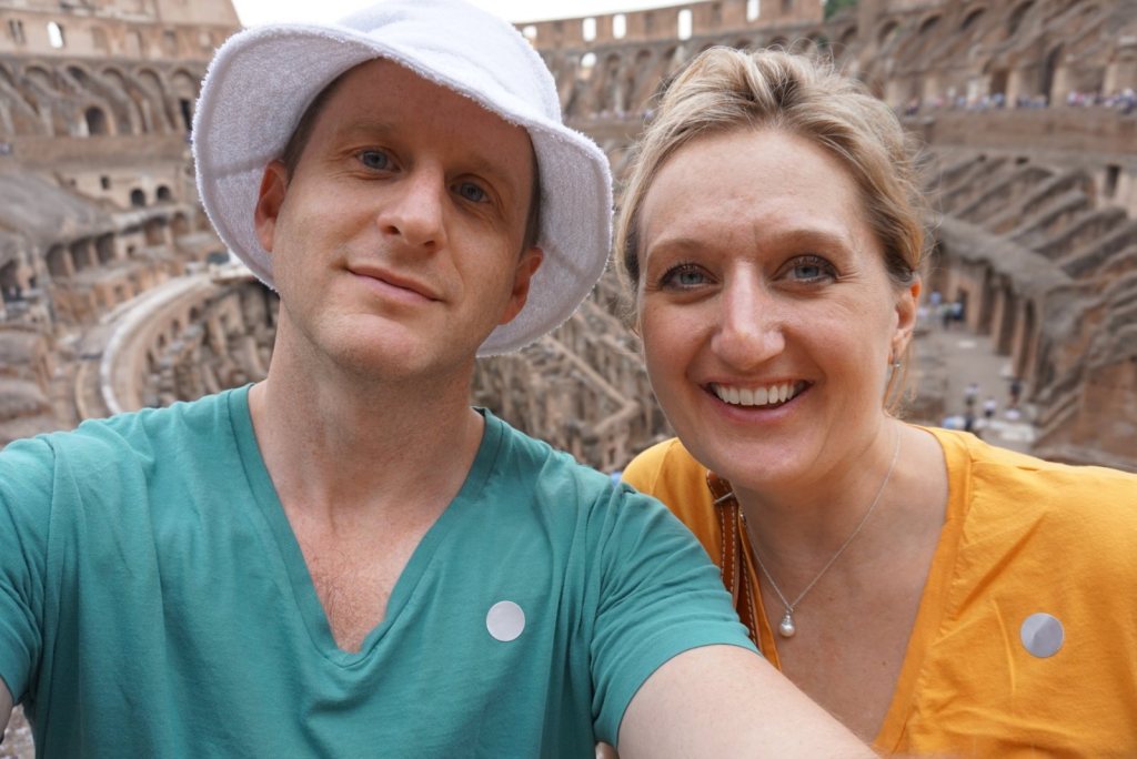 Chris in a white bucket hat and Jen in orange, the Colosseum tiers rising behind them