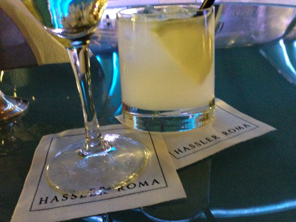 A cocktail on a paper coaster printed Hassler Roma, on a glass table in a blue-lit bar