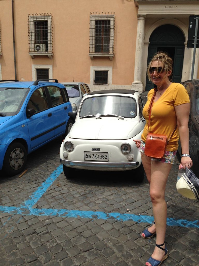 Jen beside a white Fiat 500 parked on a cobbled Rome street, a blue car behind it