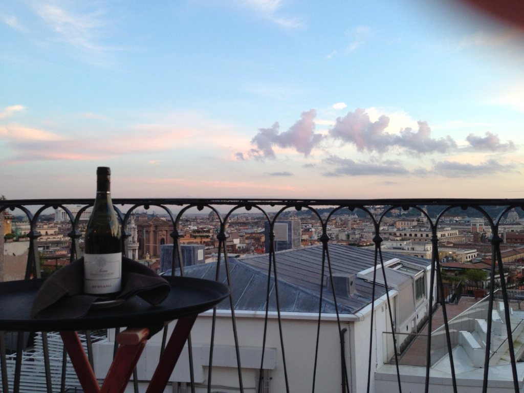 A bottle of wine on a table against the terrace ironwork, Rome's rooftops and a pink sky beyond