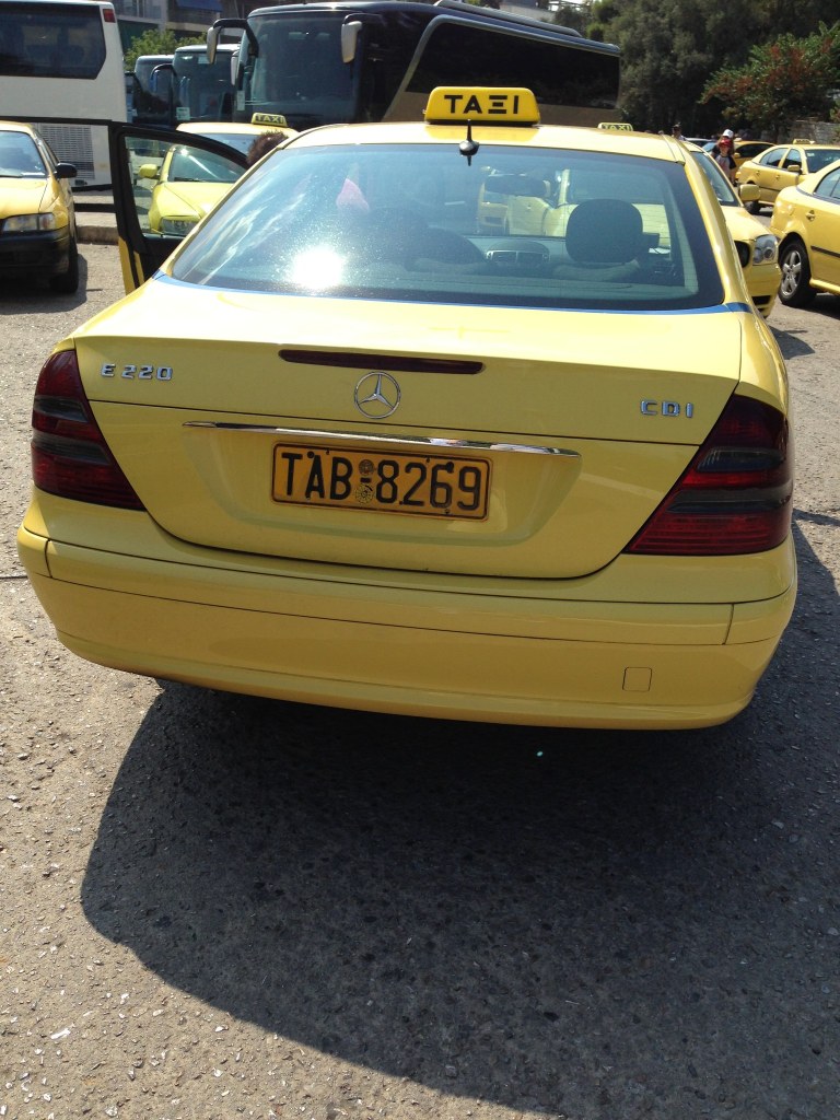 A yellow Athens taxi photographed from behind at the Acropolis rank, more taxis and two tour coaches beyond