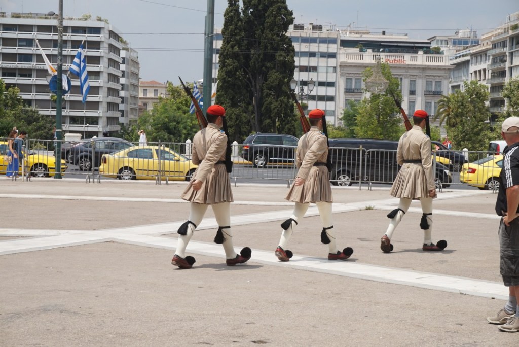 Three Evzones marching in file across the marble at Syntagma in red fezzes, pleated kilts and pom-pom shoes