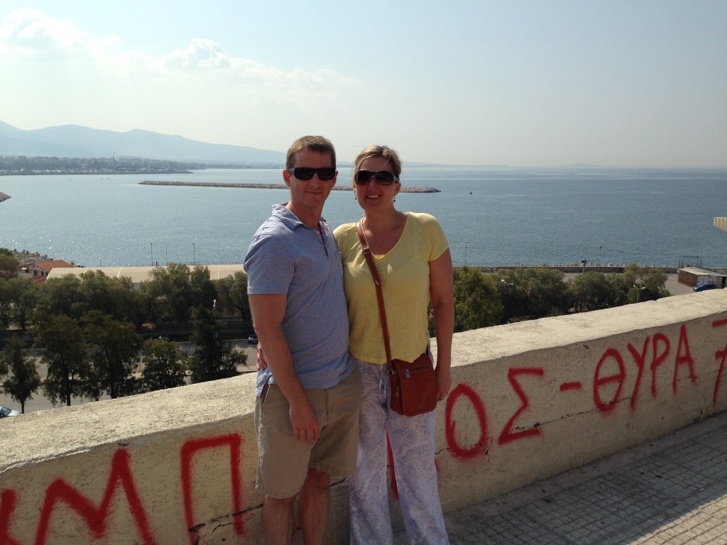 Chris and Jen on a terrace above a Piraeus marina, red Greek graffiti reading Olympiacos Gate 7 along the parapet
