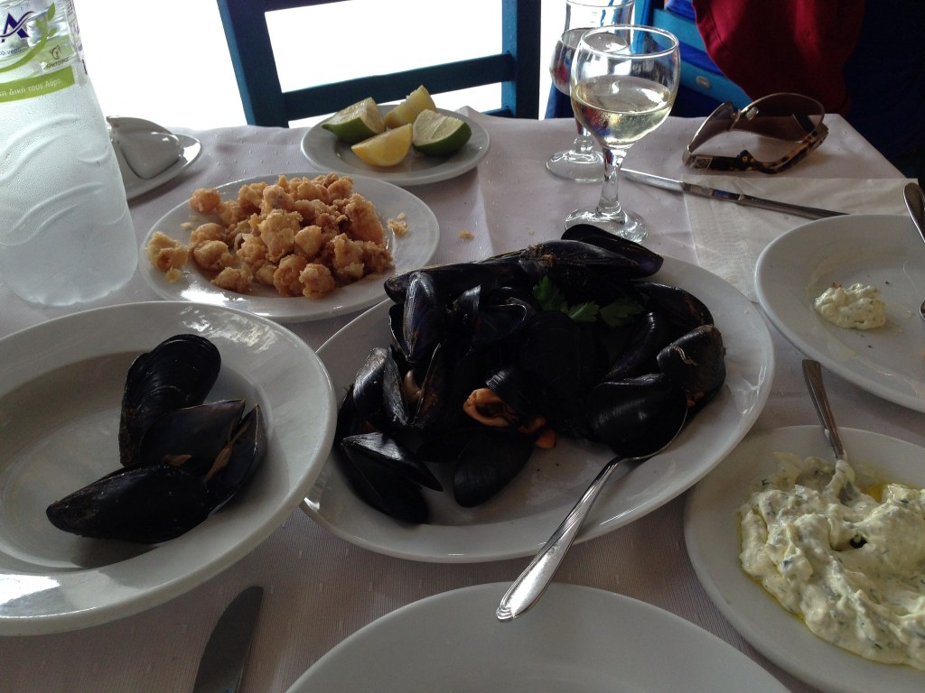 A table of mussels, small fried seafood, two plates of tzatziki, lemon and a glass of white wine