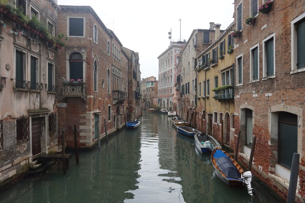 A quiet Venice canal seen from a bridge, boats moored along the wall and a gondola under the far buildings