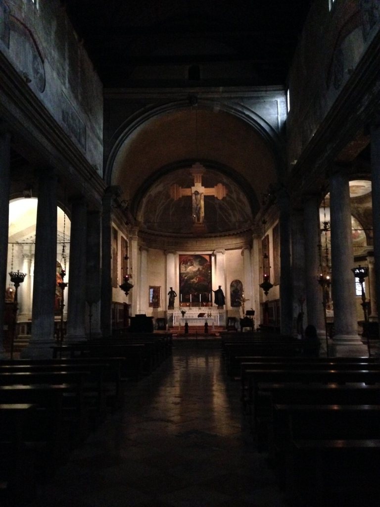 The dark interior of a Venetian parish church, columns down both sides and a painted crucifix hanging above the chancel