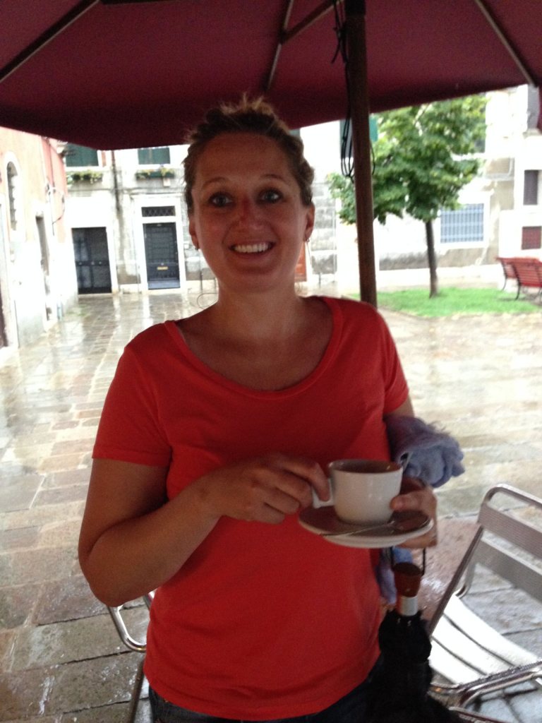 Jen holding an espresso cup under a burgundy parasol in a wet and empty Venice campo