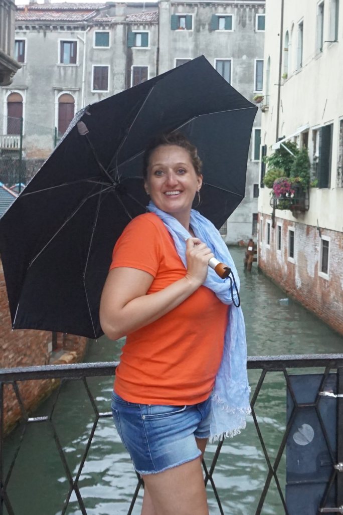 Jen under a black umbrella on an iron-railed bridge over a Venice back canal, palazzi rising on both sides