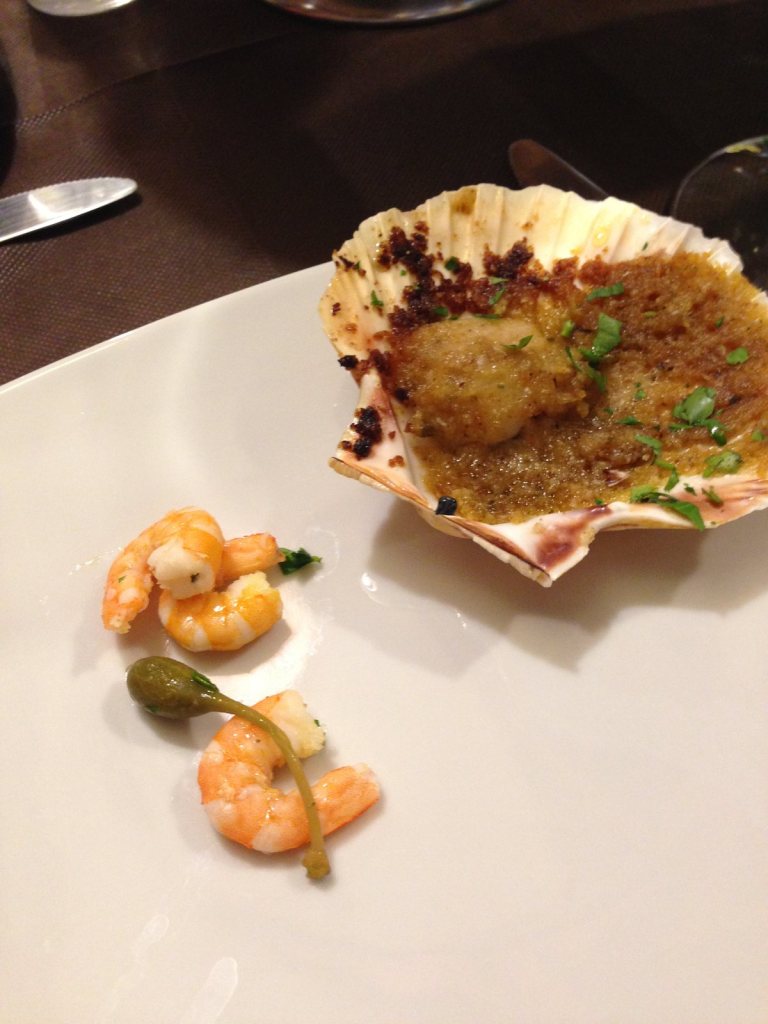 A scallop baked in its shell under breadcrumbs, three prawns and a caper on the plate beside it