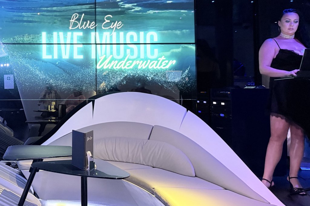 A singer in a black dress performing beside a screen reading Blue Eye Live Music Underwater, in the curved white lounge below the waterline aboard Le Bougainville.