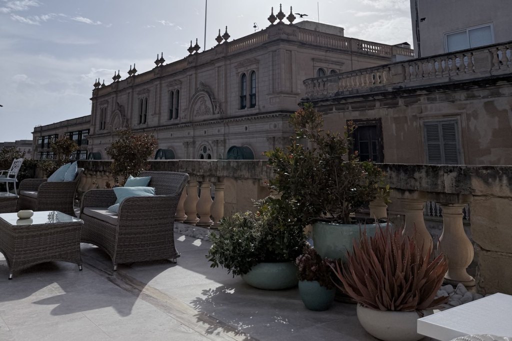 A sunlit rooftop terrace with wicker seating and potted plants, overlooking Baroque limestone rooftops in Valletta.