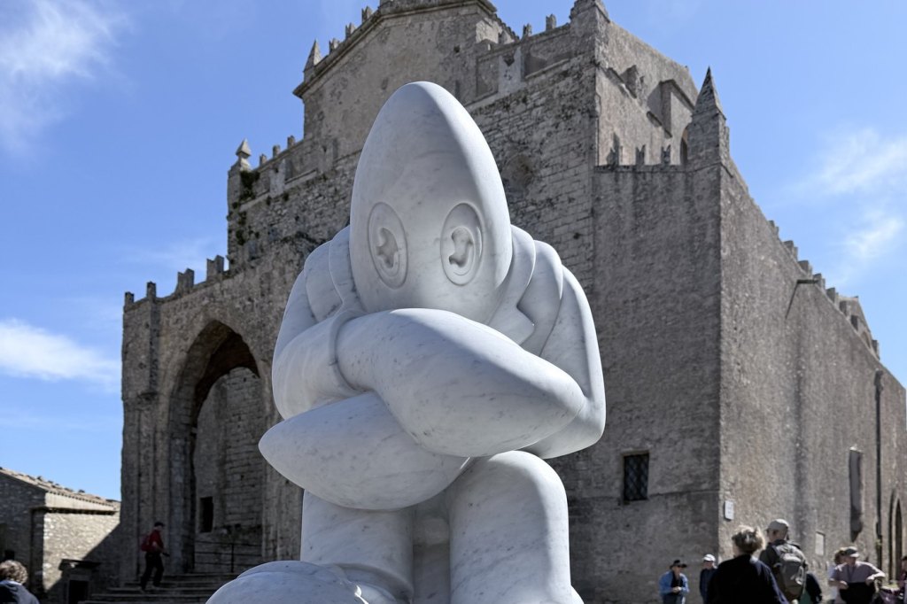 A large white marble cartoon sculpture by Philip Colbert on a plinth outside the medieval Chiesa Madre in Erice.