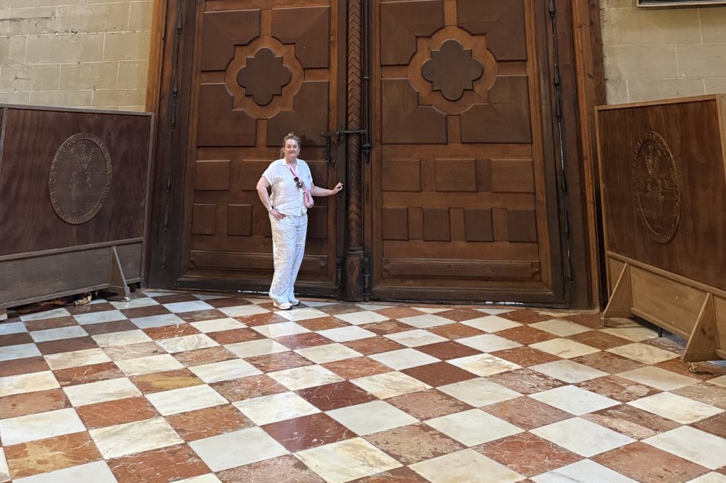 A visitor dwarfed by the towering carved wooden doors of Málaga Cathedral, on a red and white chequered marble floor.