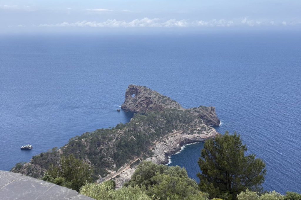 A rocky pine-covered peninsula jutting into deep blue Mediterranean water, seen from a clifftop above Deià, Mallorca.
