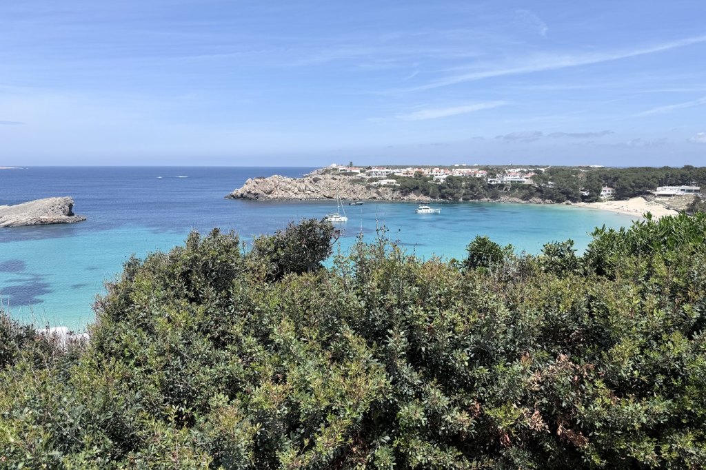 A turquoise Menorcan cove with a sandy beach and anchored sailboats, seen over Mediterranean scrubland.