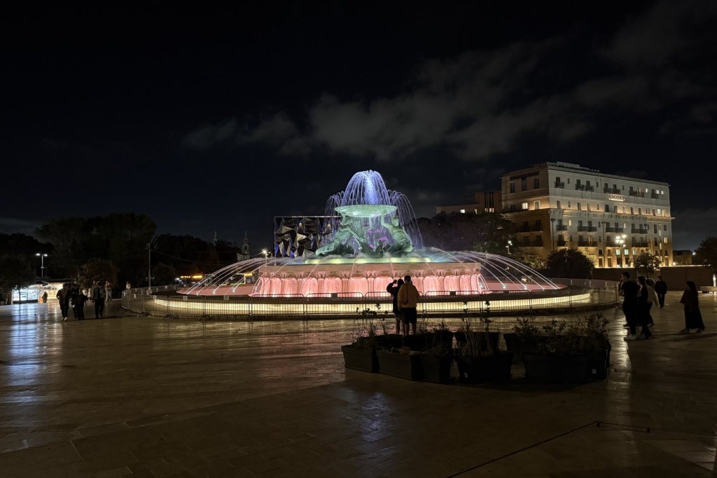 The Triton Fountain lit pink and blue at night in the City Gate plaza, Valletta.