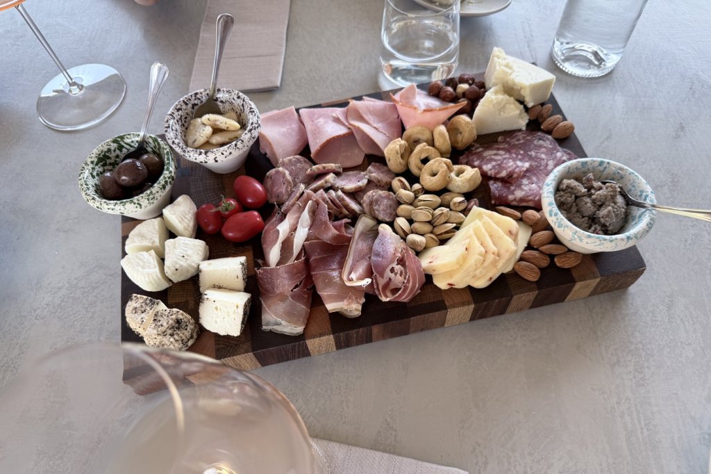 A Maltese grazing board of local cheeses, cured meats, olives and nuts beside wine glasses.