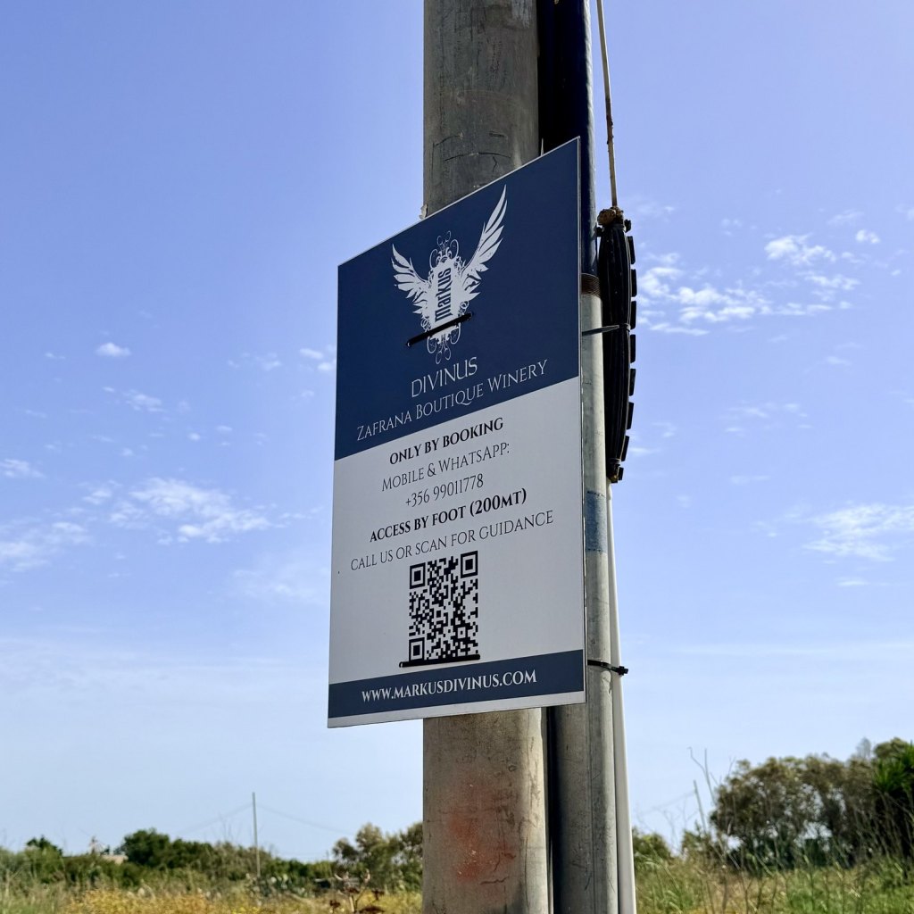 A dark blue sign cable-tied to a utility pole reading Divinus, Zafrana Boutique Winery, only by booking, access by foot 200 metres, with a QR code beneath and open scrub behind