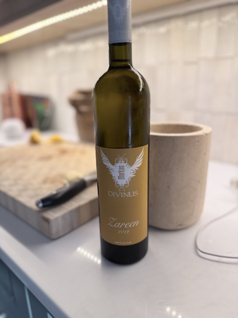 The Markus Divinus Zareen 2019 bottle standing on a kitchen counter, its gold label lit from the side, a chopping board and knife behind it