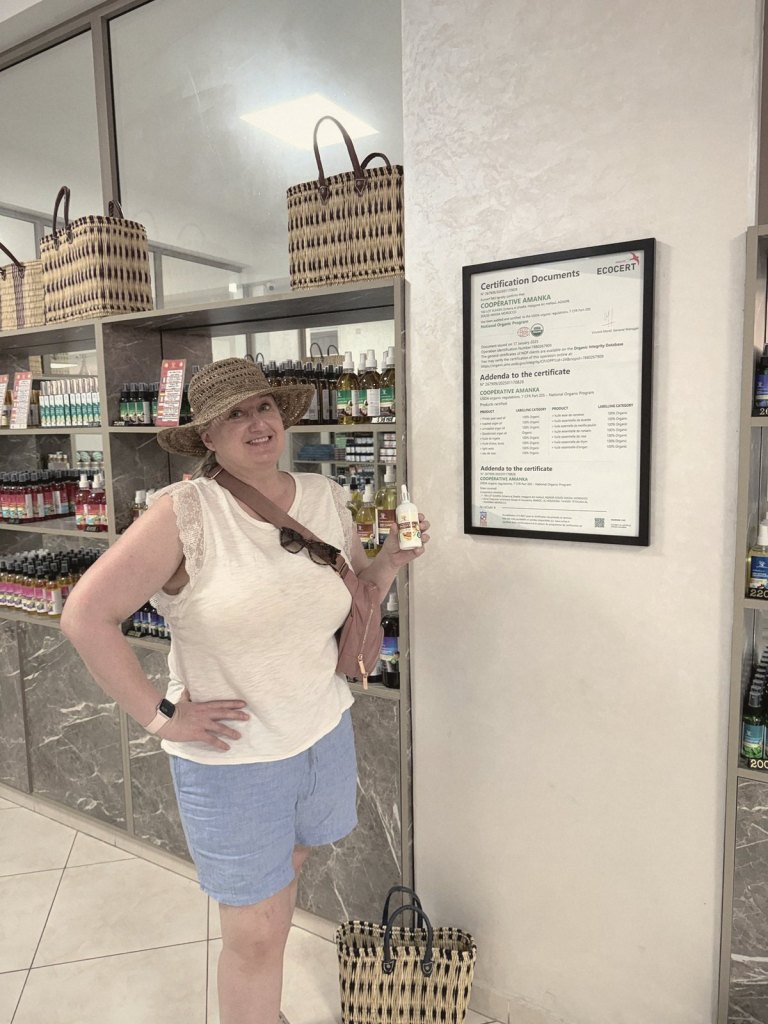 Jen in the argan cooperative's shop holding up a small bottle beside a framed certificate, with shelves of bottles and woven baskets behind her