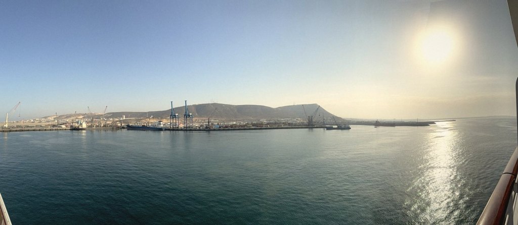 Early morning from the ship's balcony: Agadir's commercial port across calm water, cranes and quays beneath a long hill, the sun low in a hazy sky
