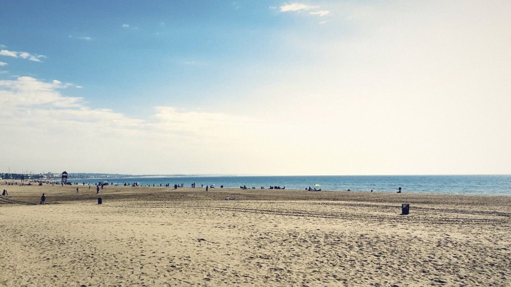 Agadir's wide sandy beach in the afternoon, a few people scattered along the waterline under a pale blue sky