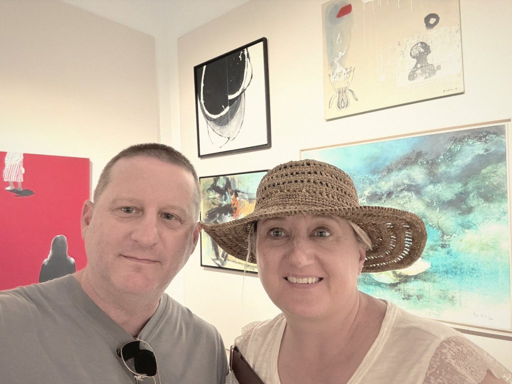 Chris and Jen, Jen in a straw hat, taking a selfie in a small white-walled gallery hung with abstract paintings