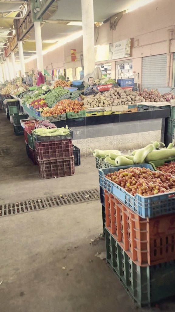 A long covered aisle in the market lined with crates of tomatoes, courgettes, peppers and potatoes under white pillars and arched shopfronts