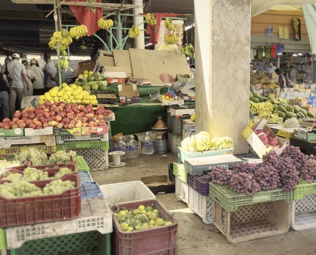 A fruit stall in the covered market: crates of green and red grapes, apples, lemons with their leaves, bananas hanging from the frame above, dragon fruit and mangoes, with shoppers in the aisle behind